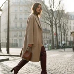 Femeie elegantă purtând cizme burgundy peste genunchi și un palton bej pe o stradă din Paris.