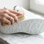 O mână curățând talpa albă a unui pantof sport cu o perie pe o suprafață de marmură, lumină naturală.
